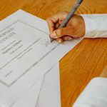 A close-up of a hand signing a child adoption certificate with a pen on a wooden table.