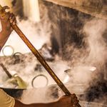 A person stirring a large pot filled with boiling liquid, surrounded by steam and heat.