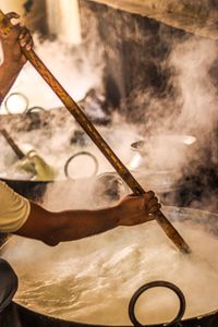 A person stirring a large pot filled with boiling liquid, surrounded by steam and heat.