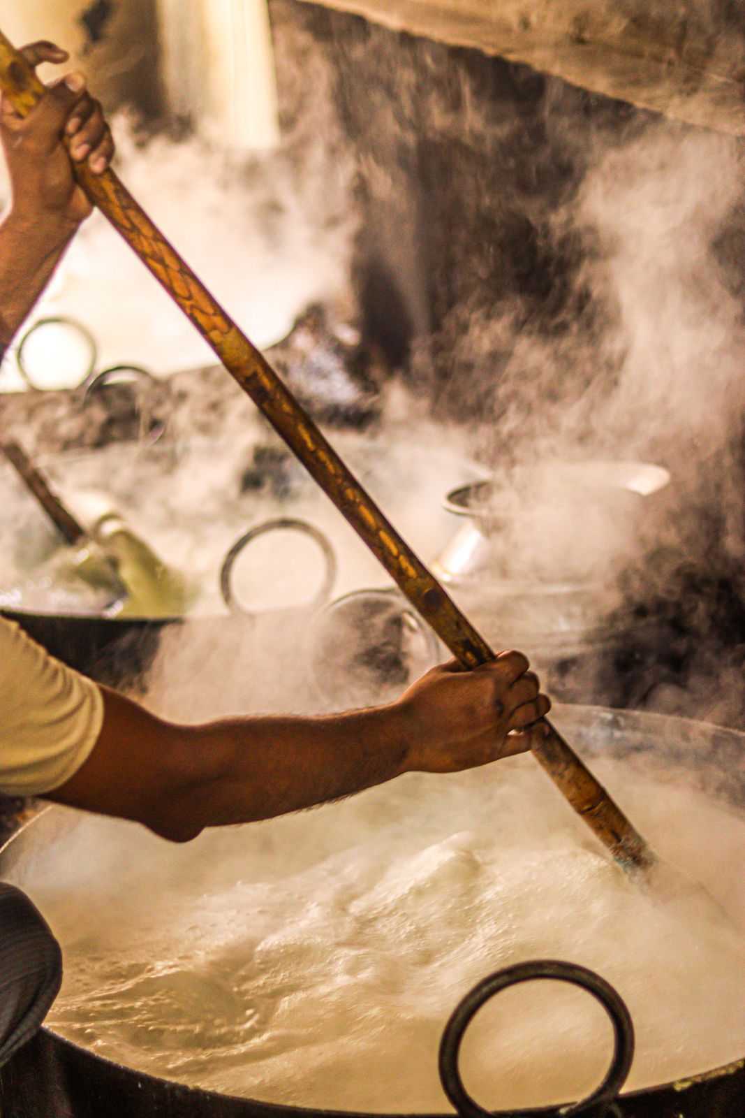 A person stirring a large pot filled with boiling liquid, surrounded by steam and heat.
