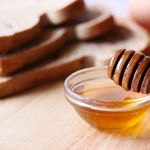 Close-up of honey dipper in a bowl with honey on wooden table beside sliced bread.
