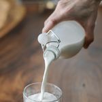 Crop anonymous person with bottle pouring milk into ornamental transparent glass on wooden table at home