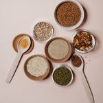 Flat lay of various grains and nuts in bowls and wooden spoons on neutral background.