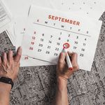 Hands holding a calendar with September dates encircled, surrounded by documents on a carpet.