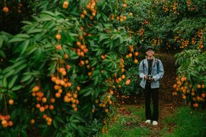 Smiling photographer standing in a vibrant lychee orchard holding a camera.