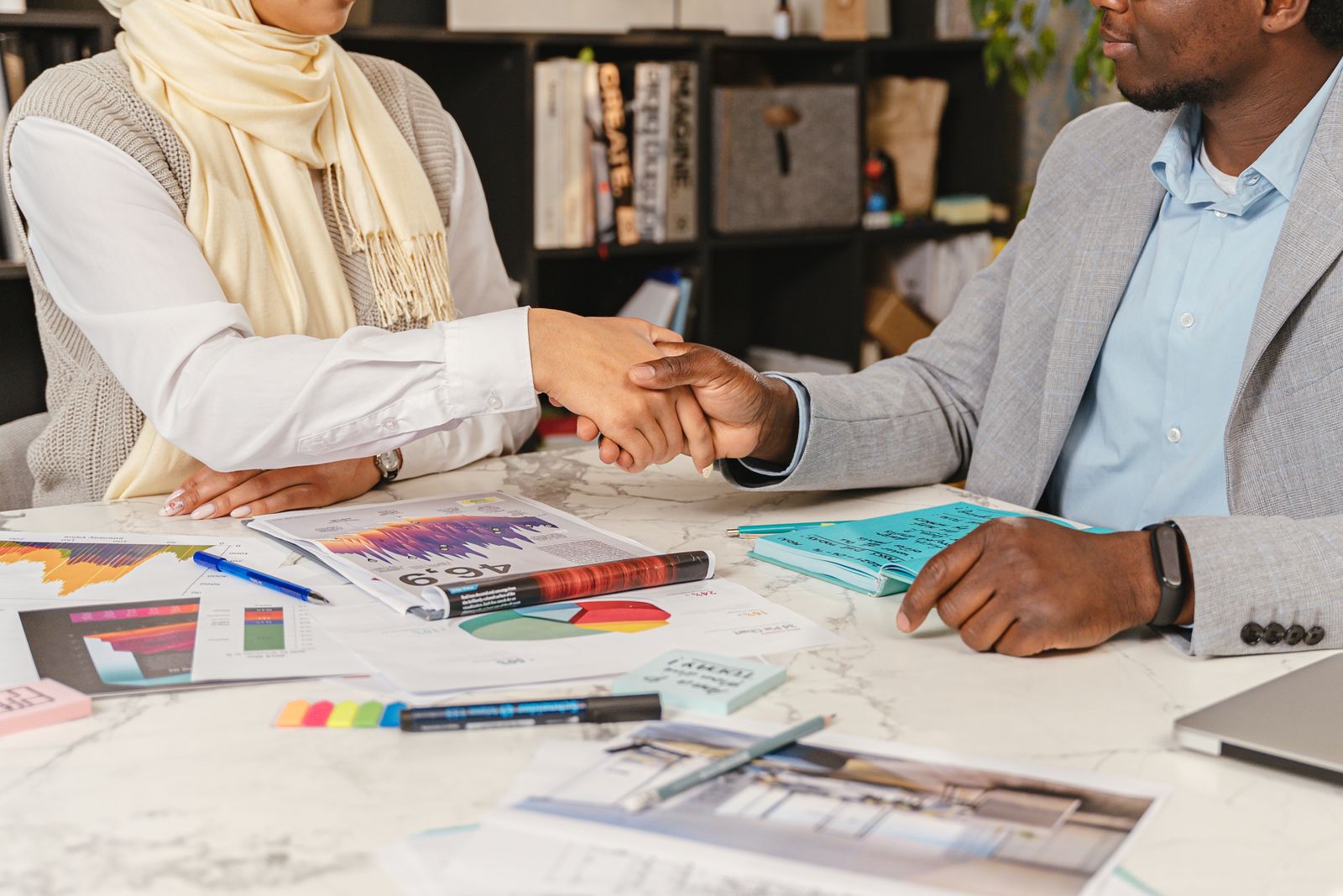 Two business professionals shaking hands over financial documents in an office setting.