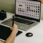 Woman using pen tablet with laptop and camera in creative workspace.