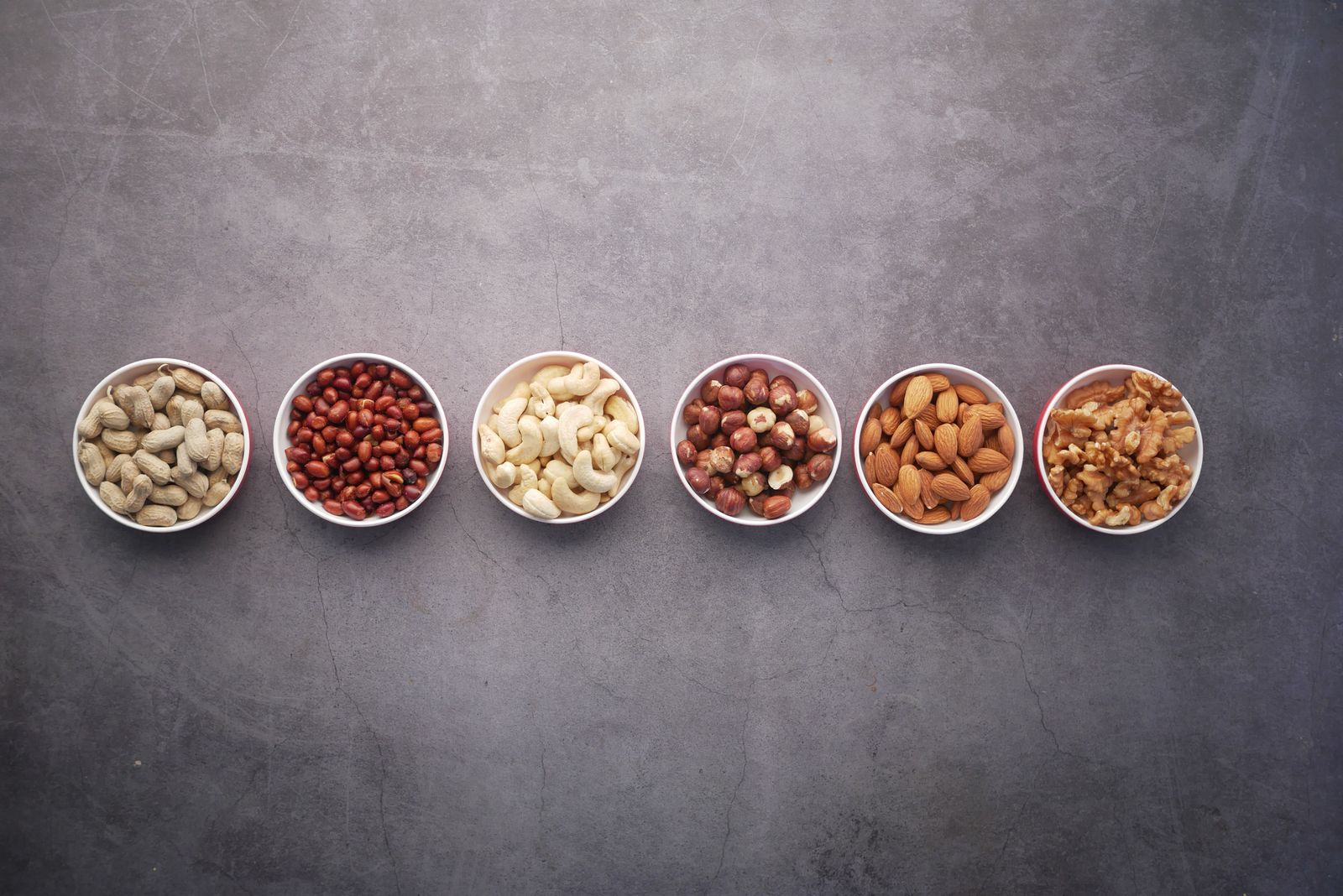 Flat lay of assorted nuts in bowls on a textured gray surface, highlighting healthy food options.